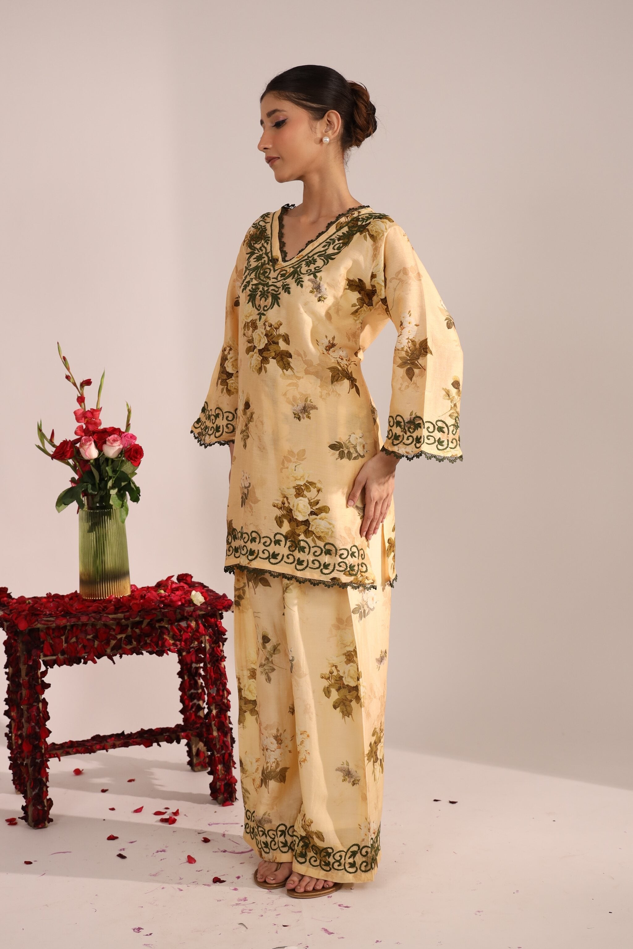 Woman wearing a floral bege chikankari co ord set in premium chaderi fabri for women standing next to a table with flowers on a light background