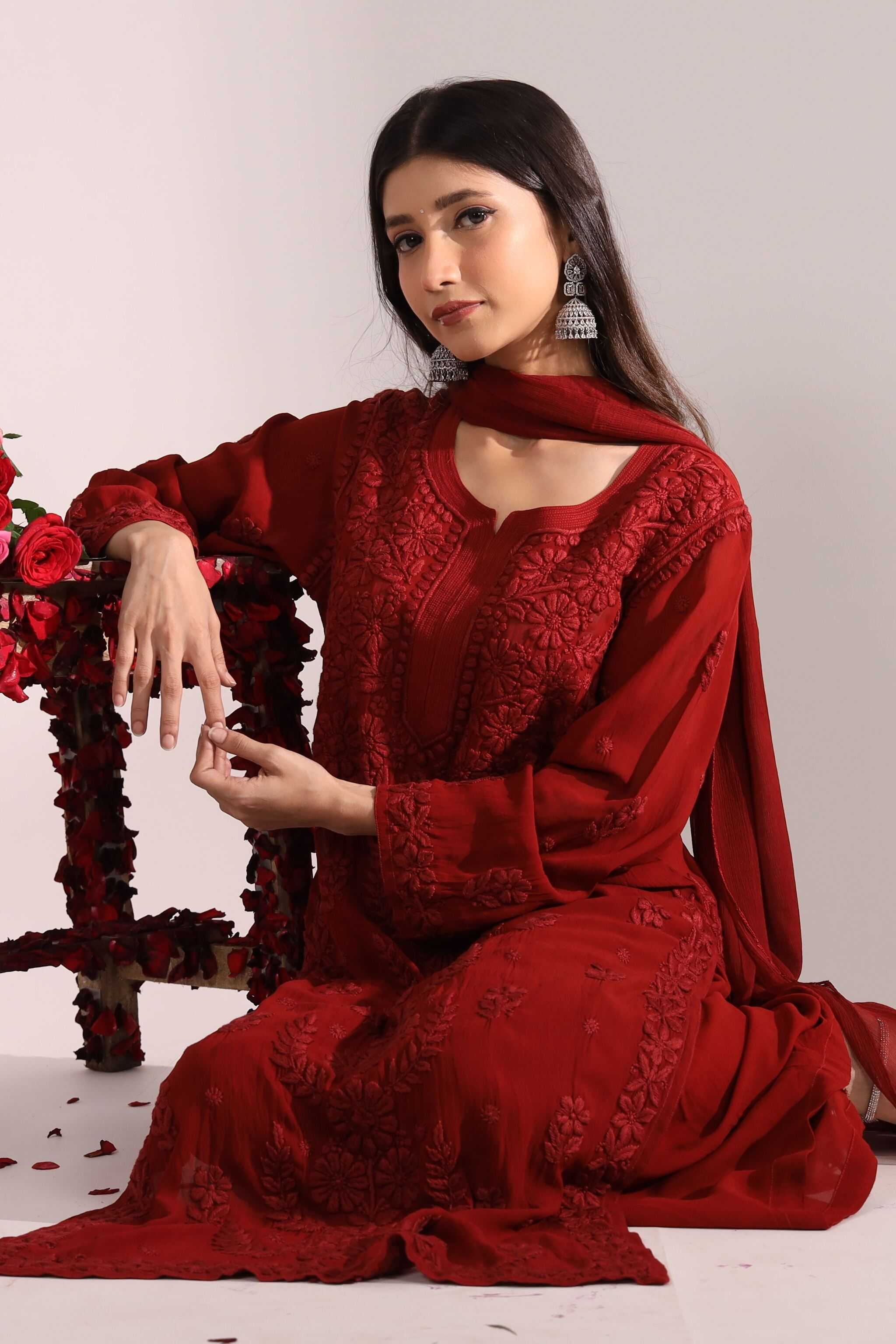 Woman wearing a red embroidered Naina chikankari bridal suit sitting on a floor with flowers.