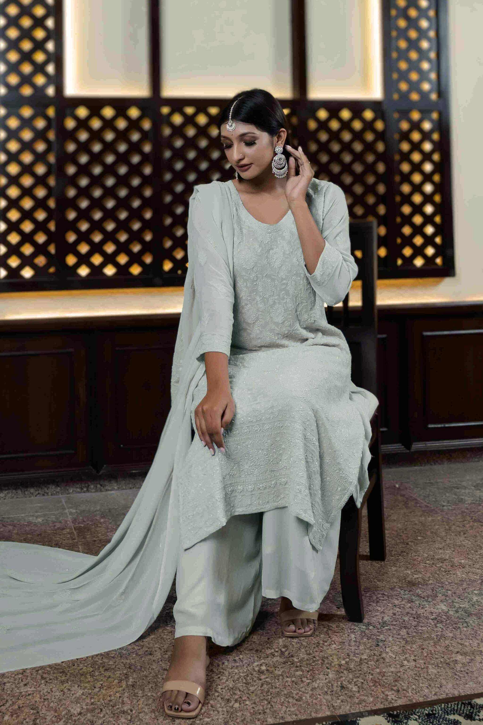 Woman in a light gray traditional Chaand Georgette Chikankari Kurta Set With Dupatta sitting on a chair with a decorative wall in the background.