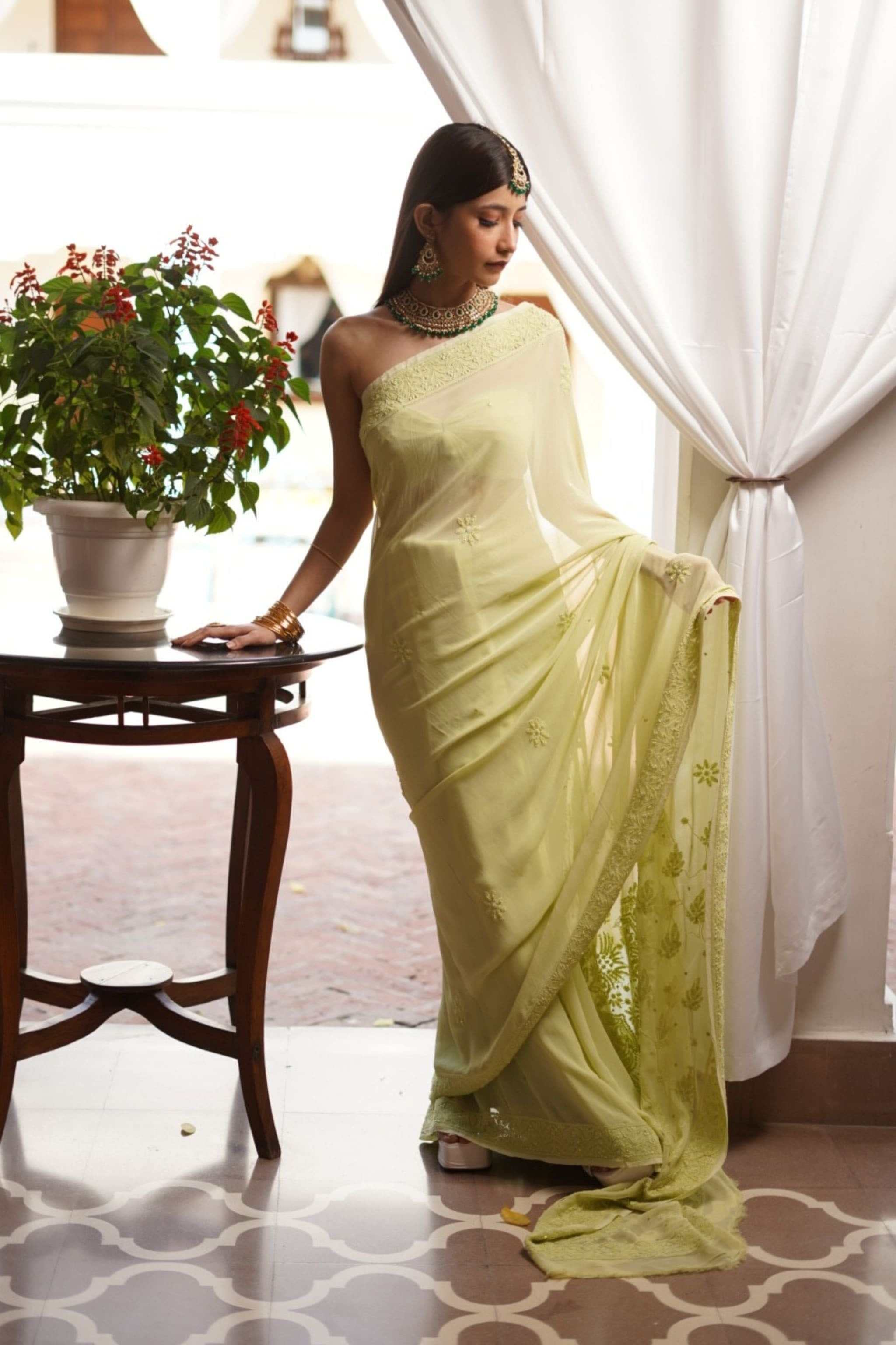 Woman in a light green chikankari saree standing indoors next to a table with a plant.