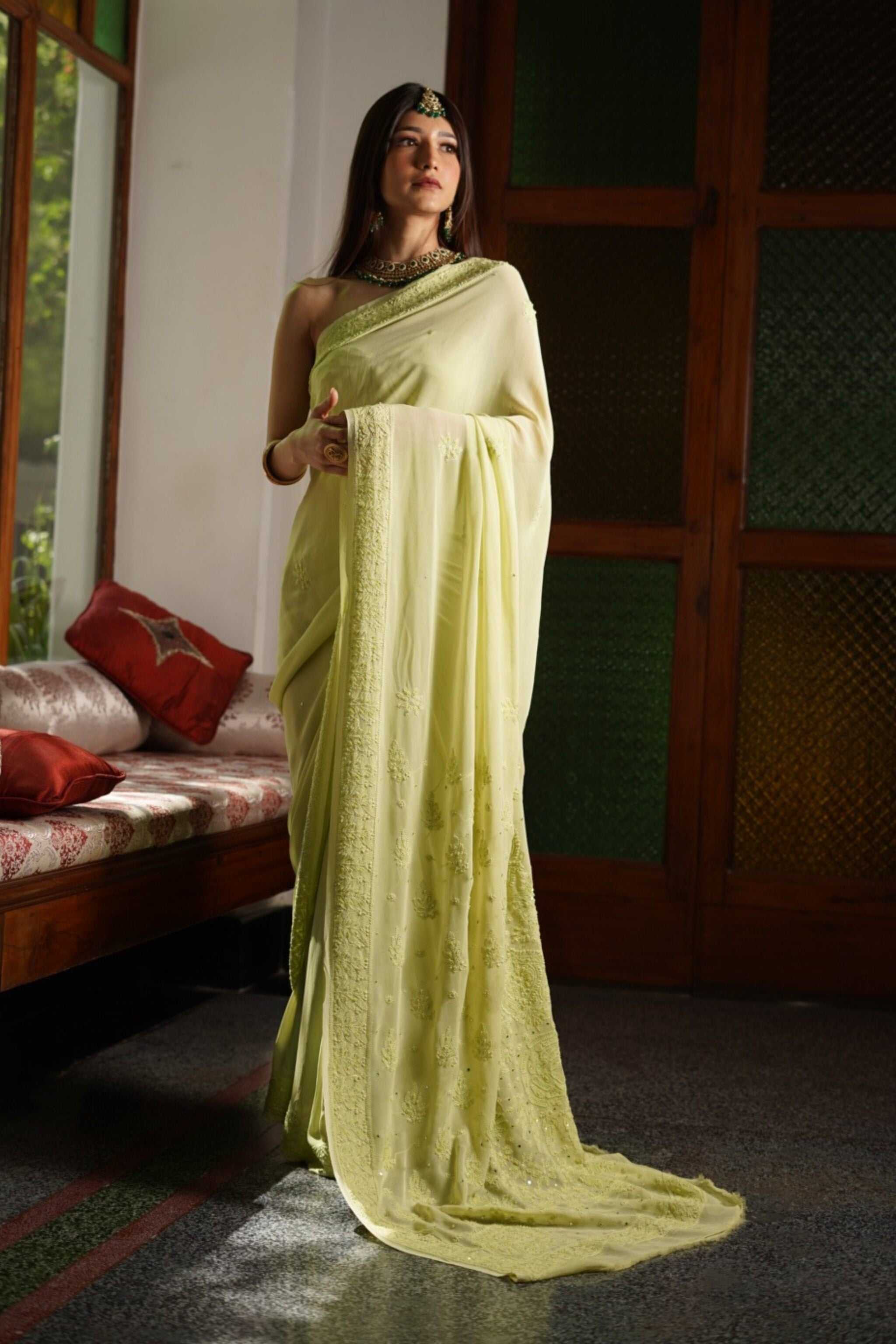 Woman in a light green chikankari saree standing indoors with a wooden door and red pillows in the background.
