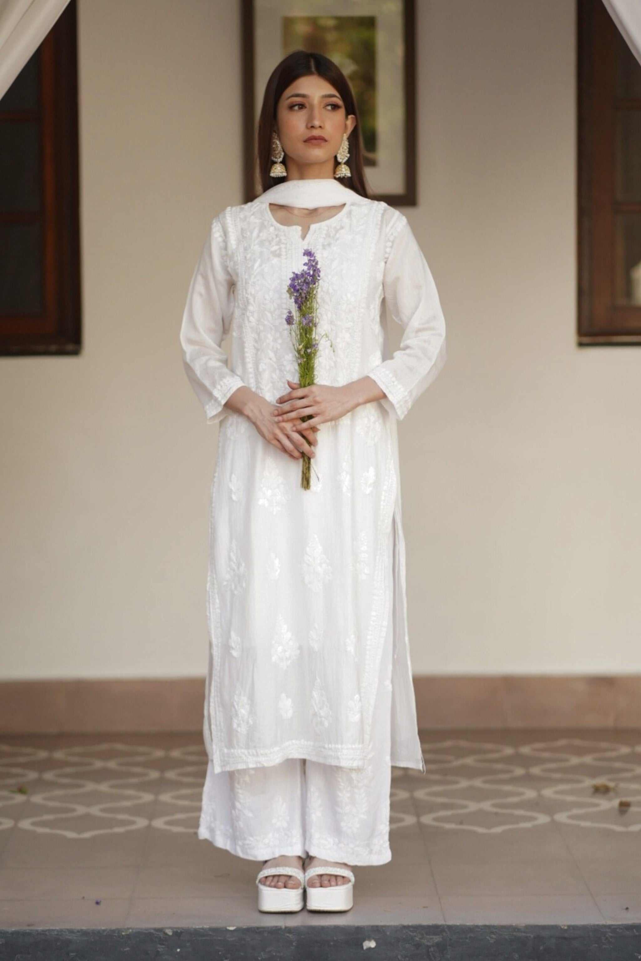 Woman in a white traditional chikankari chanderi kurta set holding flowers indoors.