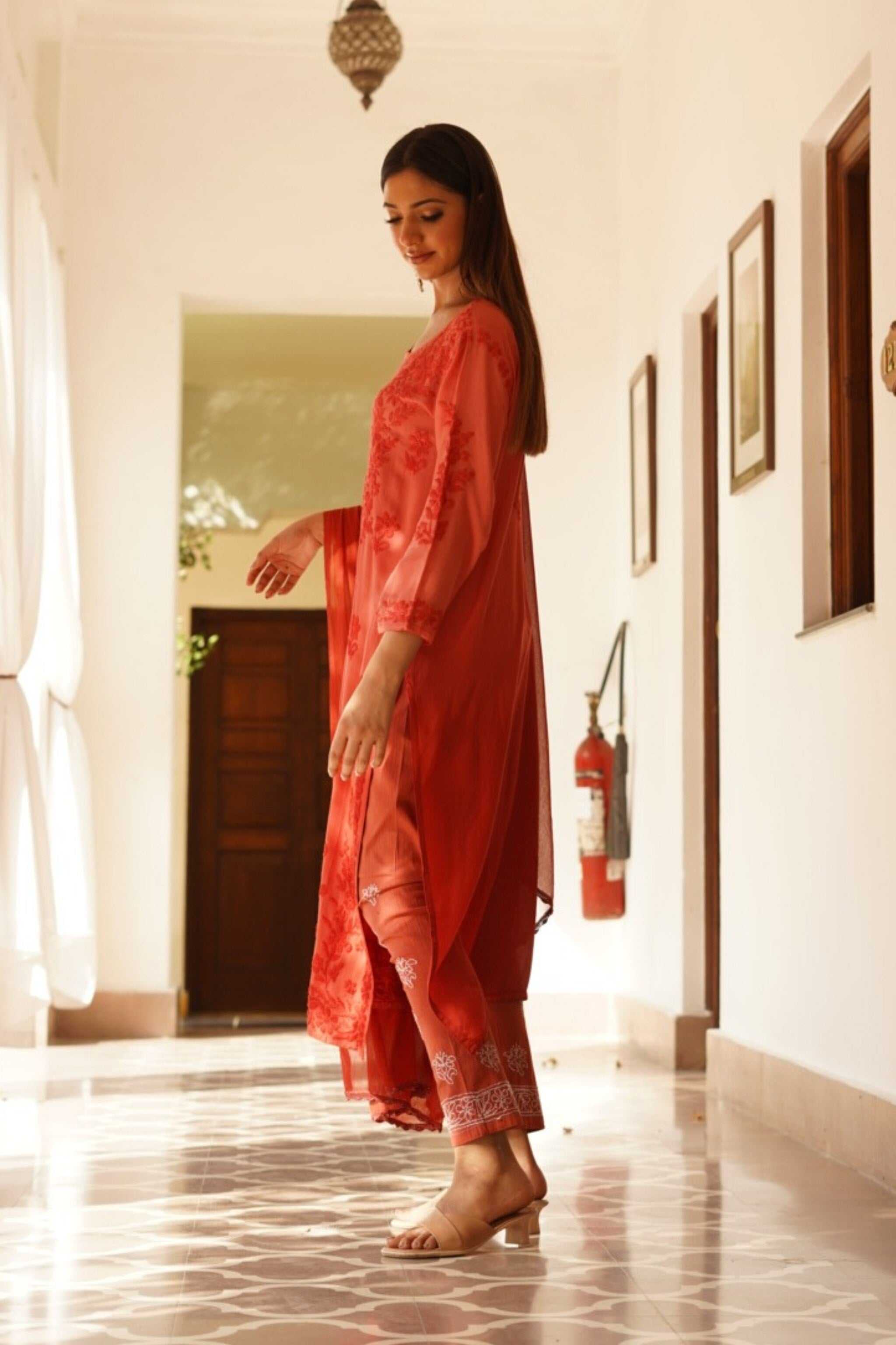Woman in a red chikan kurta set standing in a hallway with white walls and a door.