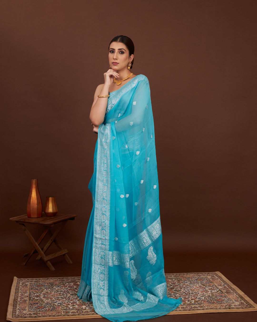 Woman wearing a turquoise saree with silver zari patterns on a brown background