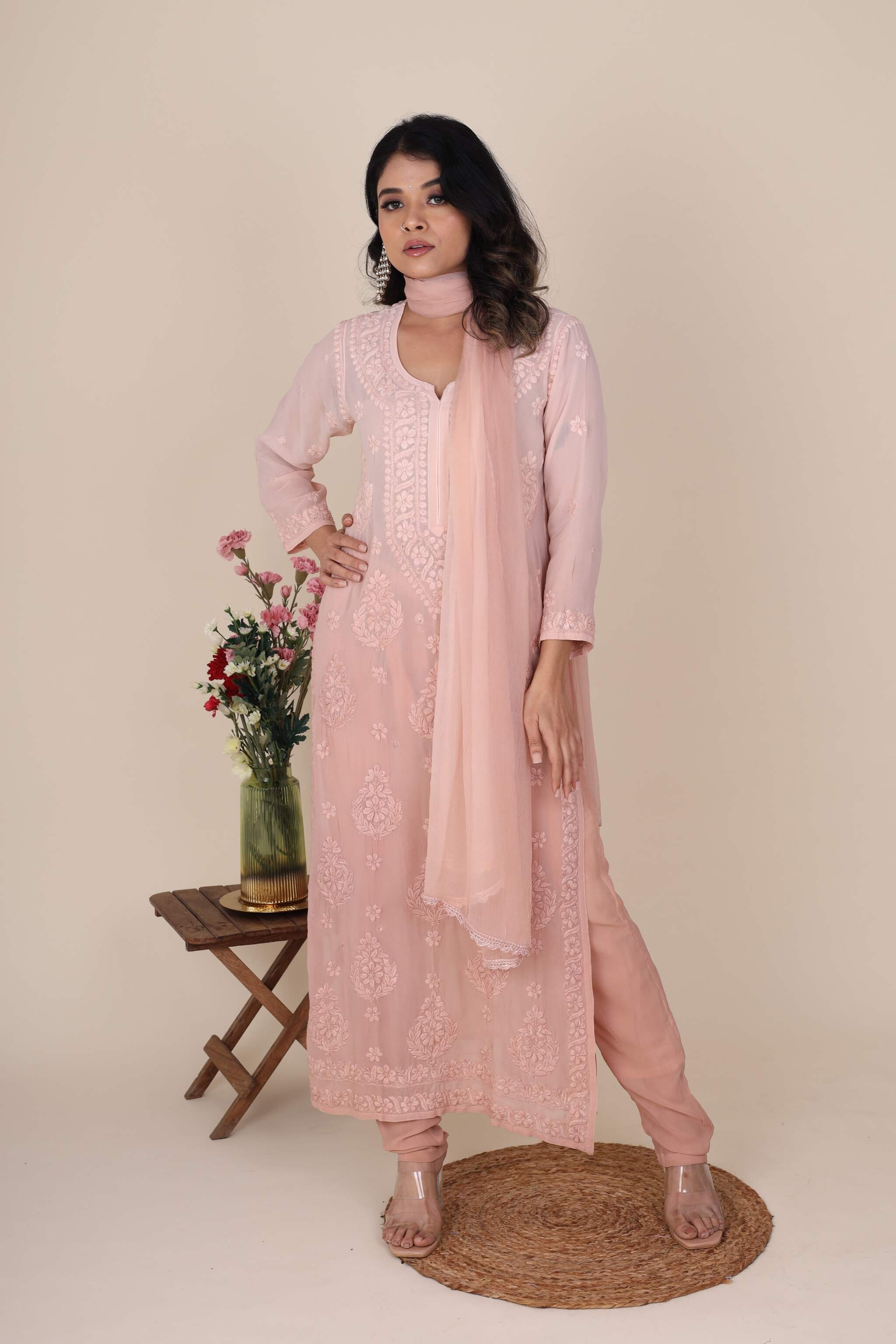 Woman wearing a pink traditional chikankari heer rose quartz set with a matching dupatta, standing next to a small table with flowers on a beige background.