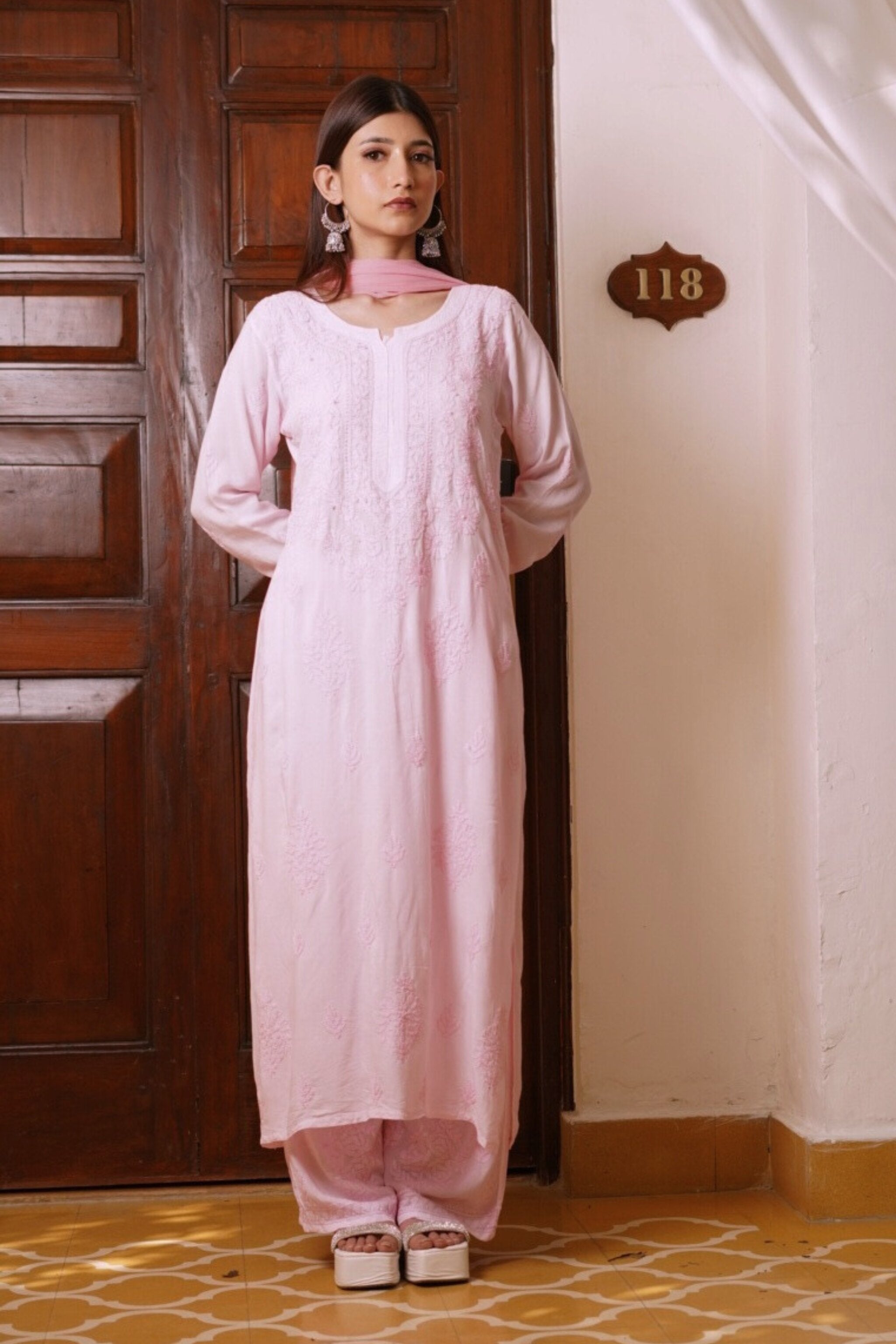 Woman wearing a light pink traditional chikan kurti set standing in front of a wooden door.