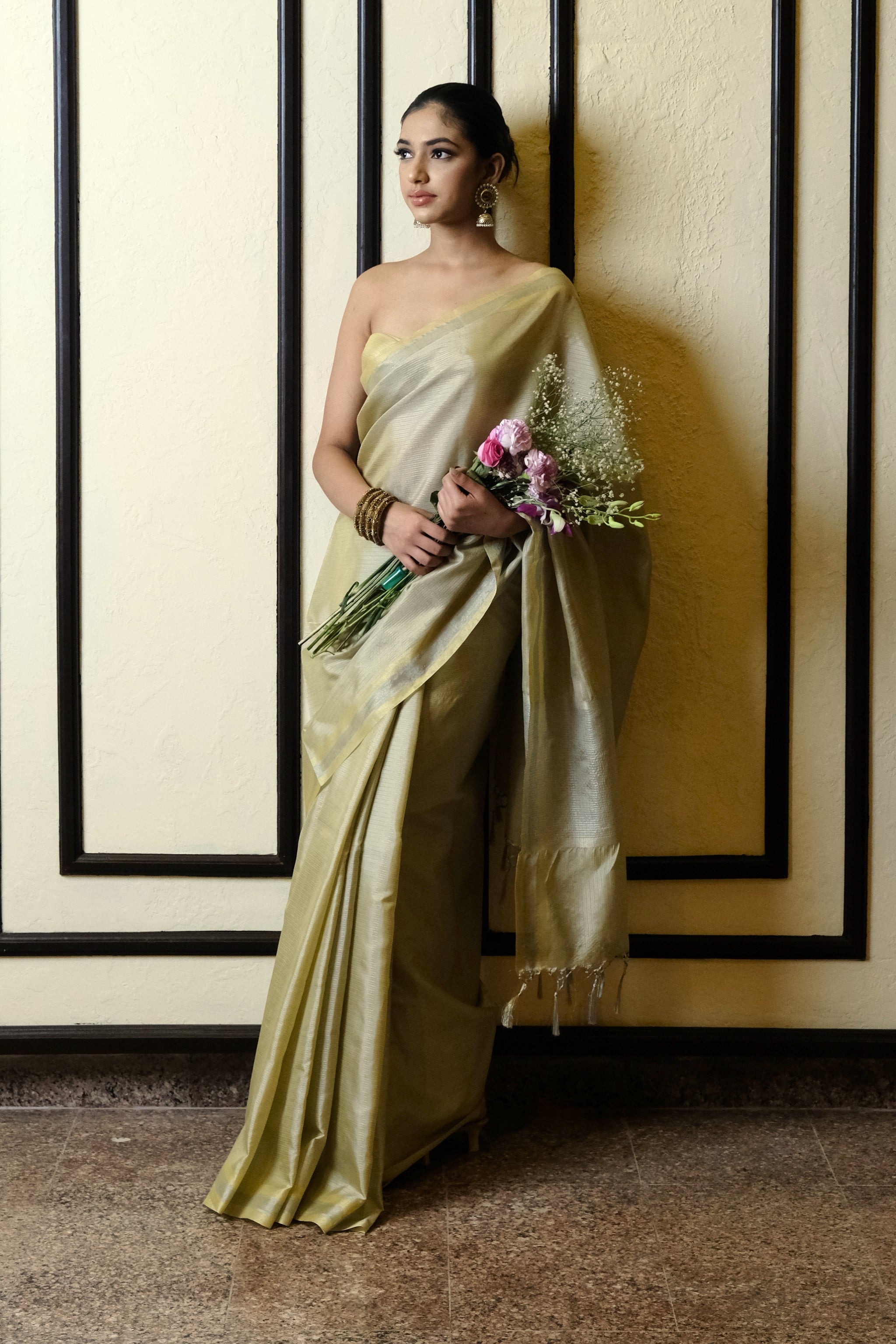 A woman wearing a sheen gold tissue banarasi saree stands against a cream-colored wall with black vertical trim, holding a bouquet of pink and white flowers.