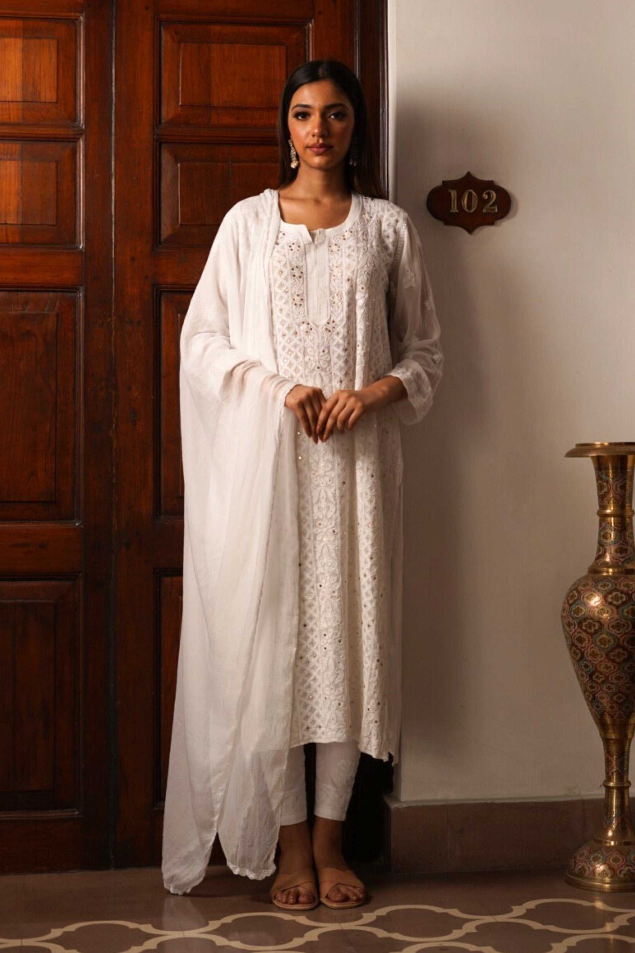 Woman in a white traditional georgette chikankari kurta set standing against a wooden door.