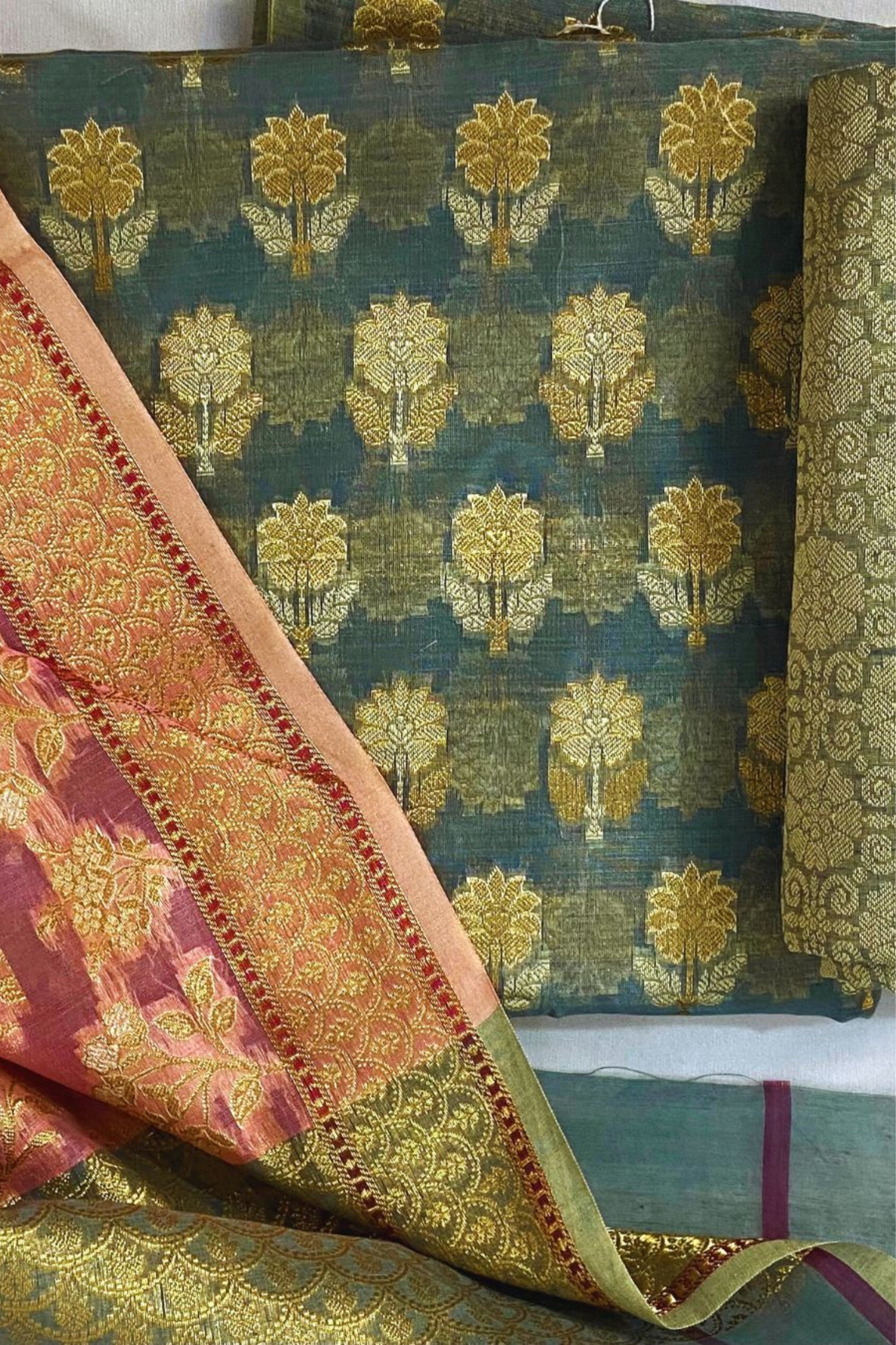 A folded and draped banarasi slk suit piece of ornate brocade fabric with a dark teal-blue base, gold floral motifs, and a contrasting pink and gold patterned bordered dupatta.
