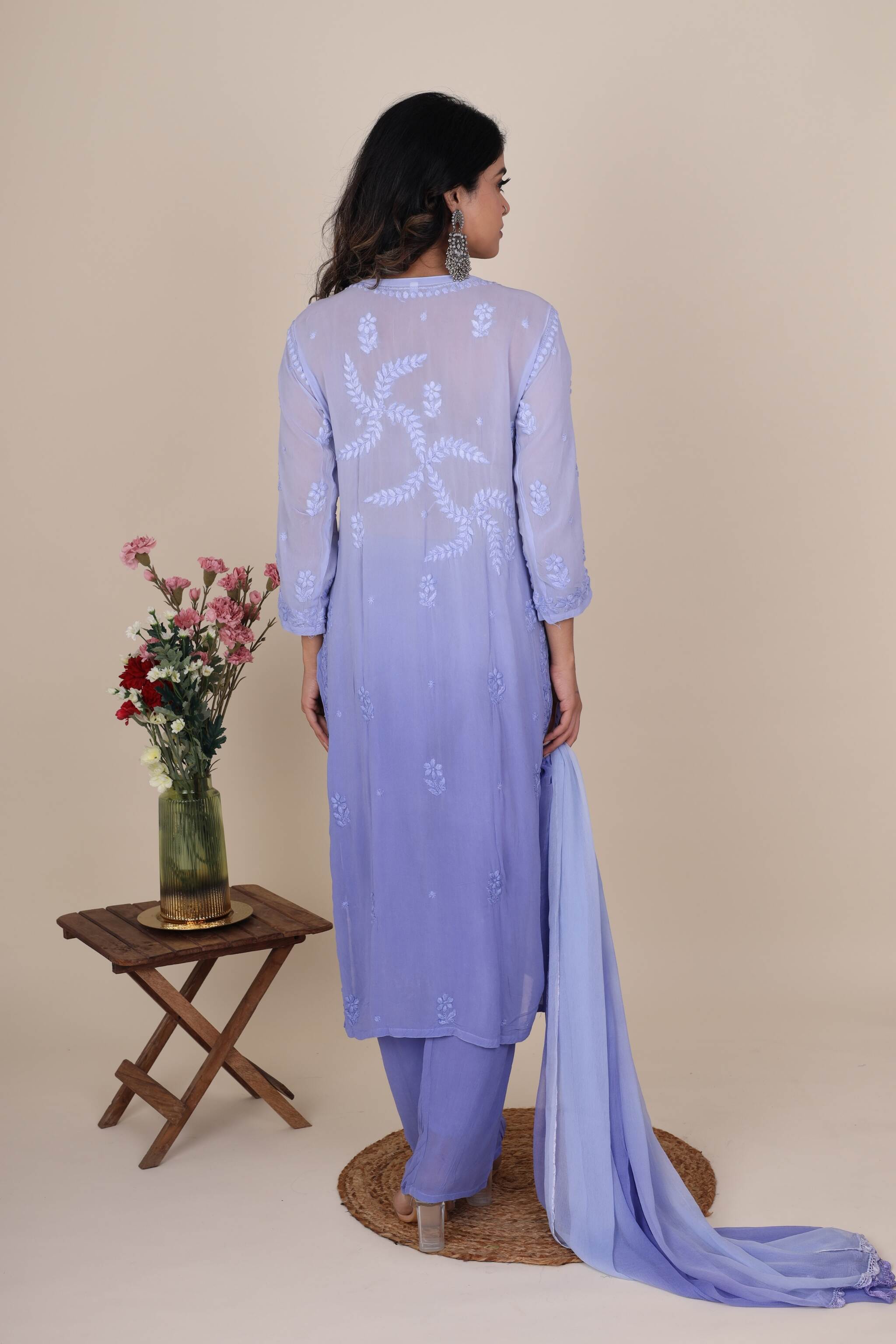 Woman wearing a light purple embroidered Chikankari Heer Topaz Set standing in front of a beige background with a small wooden table and flowers.