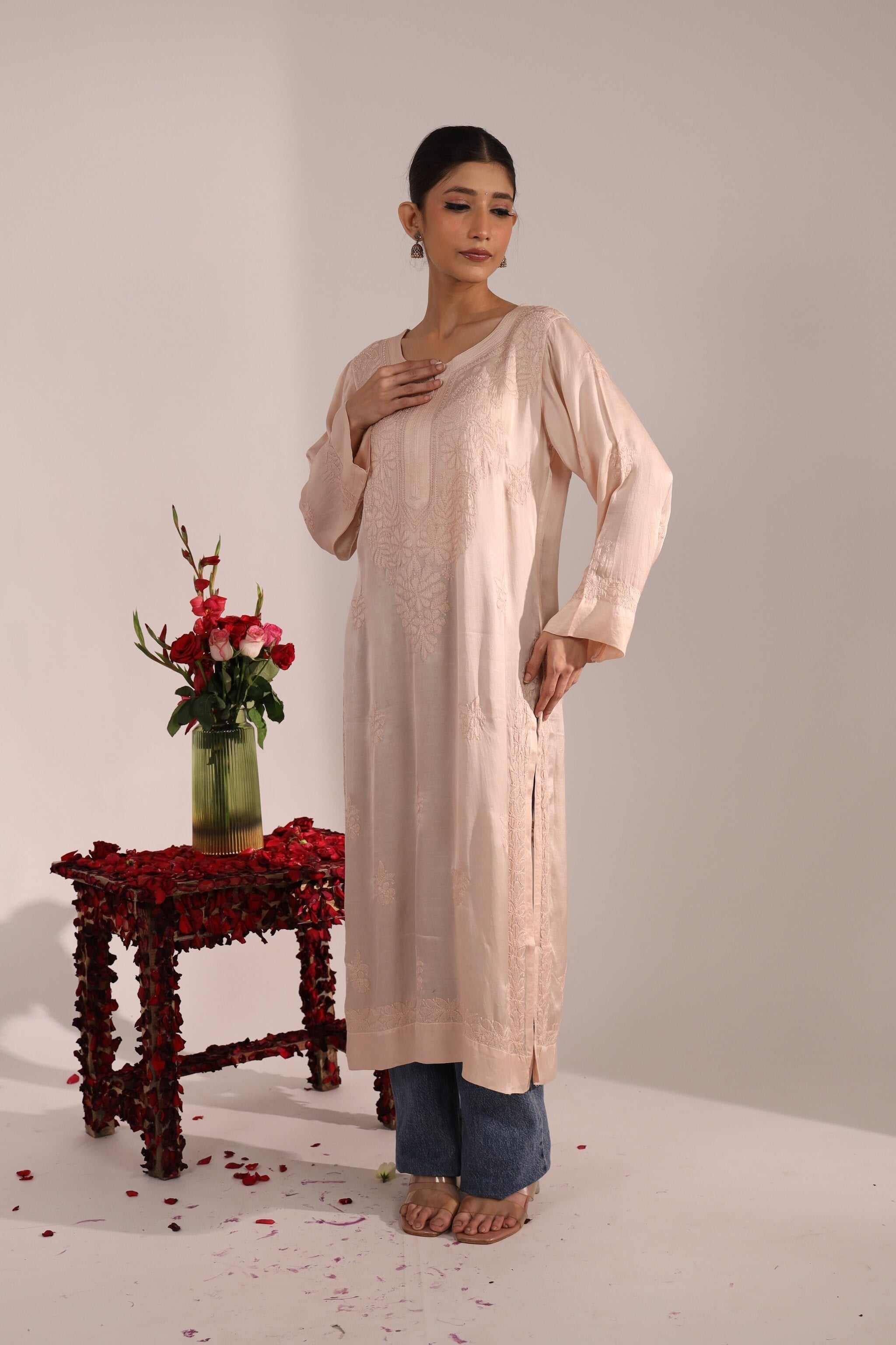 Woman wearing pastel pink Chikan Kurti paired with jeans, standing near a wooden table with flowers – elegant Indo-western look.