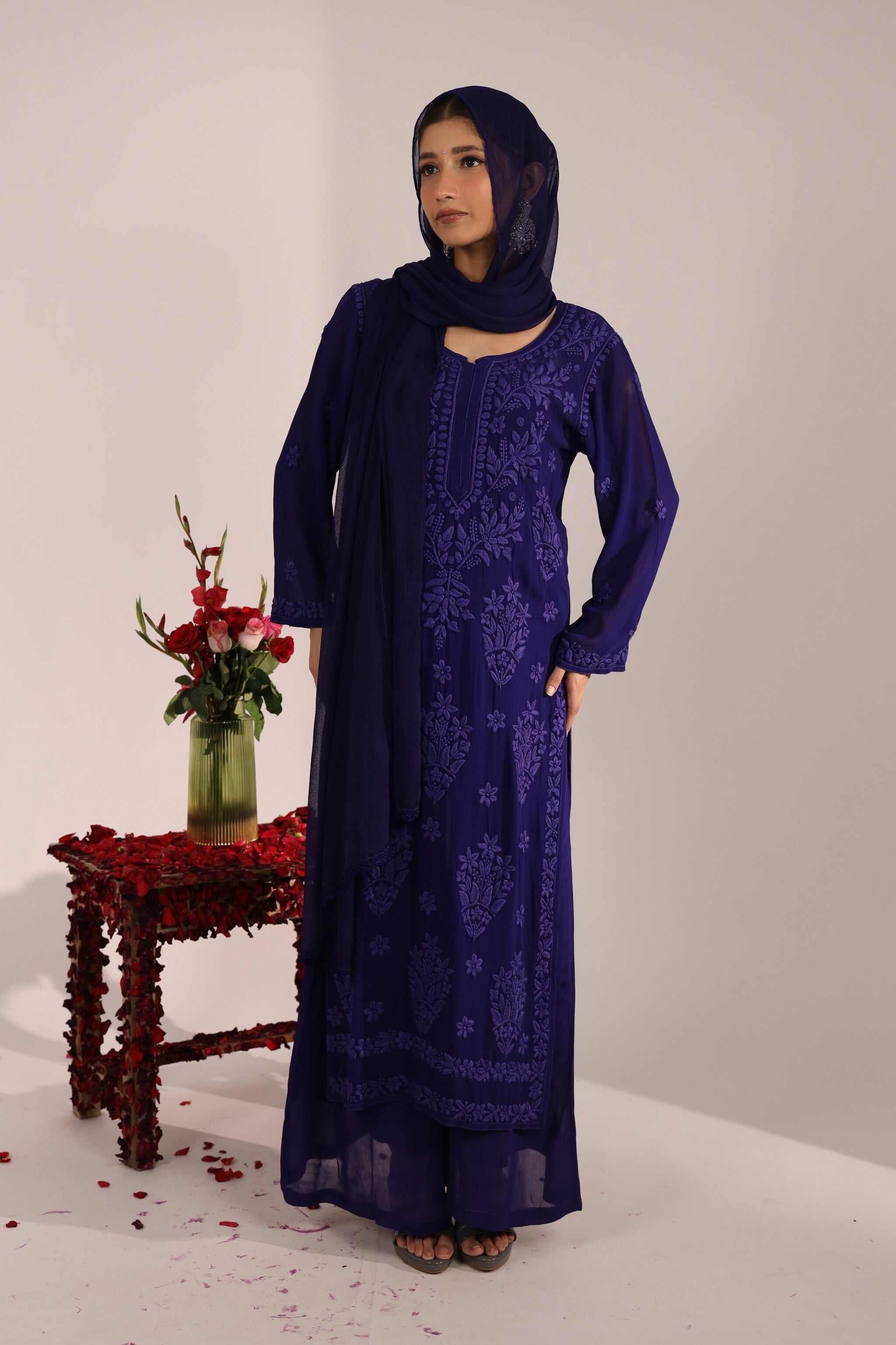 Woman wearing a blue embroidered Nayantara chikan suit and chiffon dupatta standing next to a small table with flowers on a light background