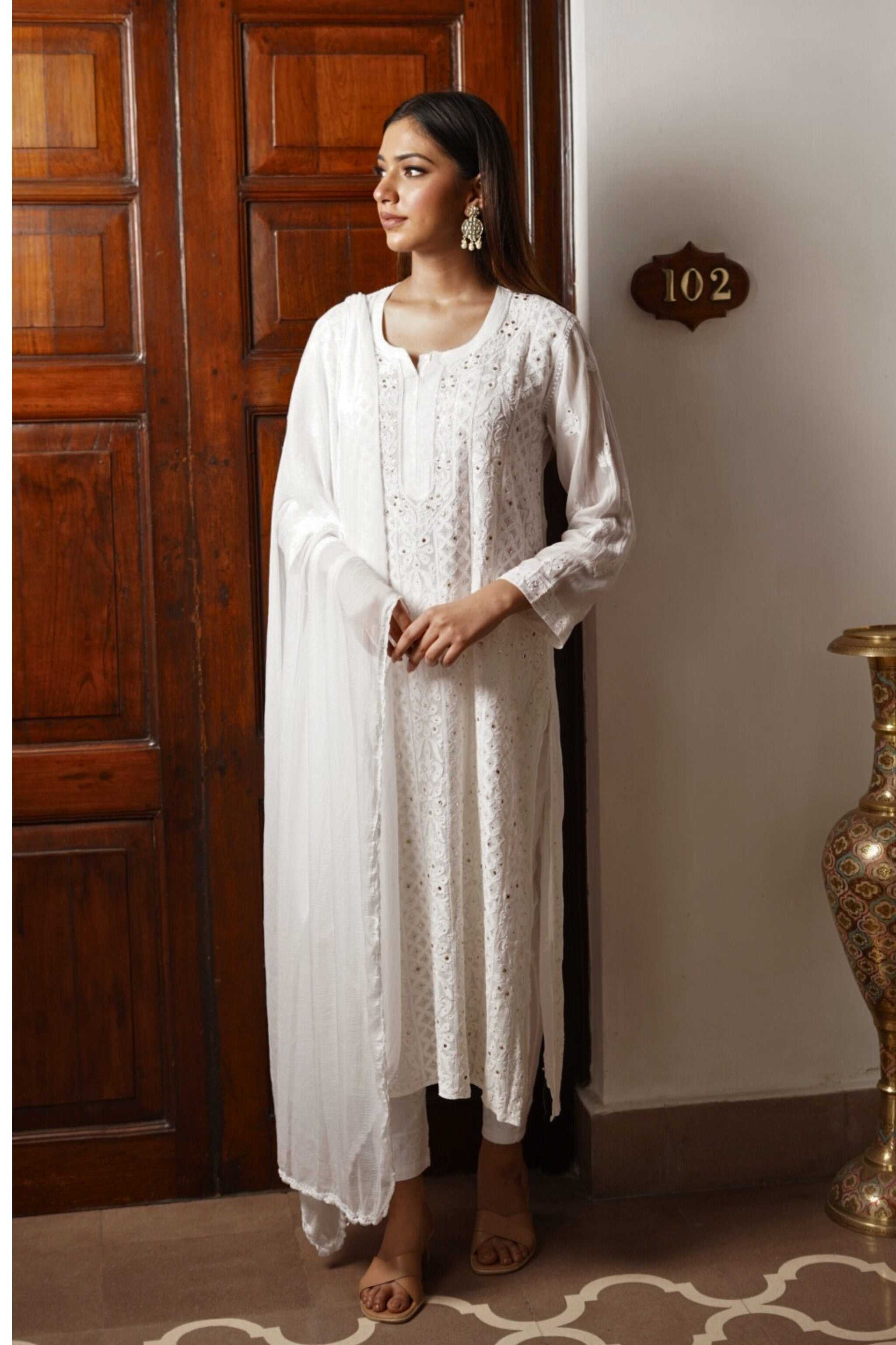 Woman in a white traditional chikankari suit standing in a room with wooden door and decorative wall.