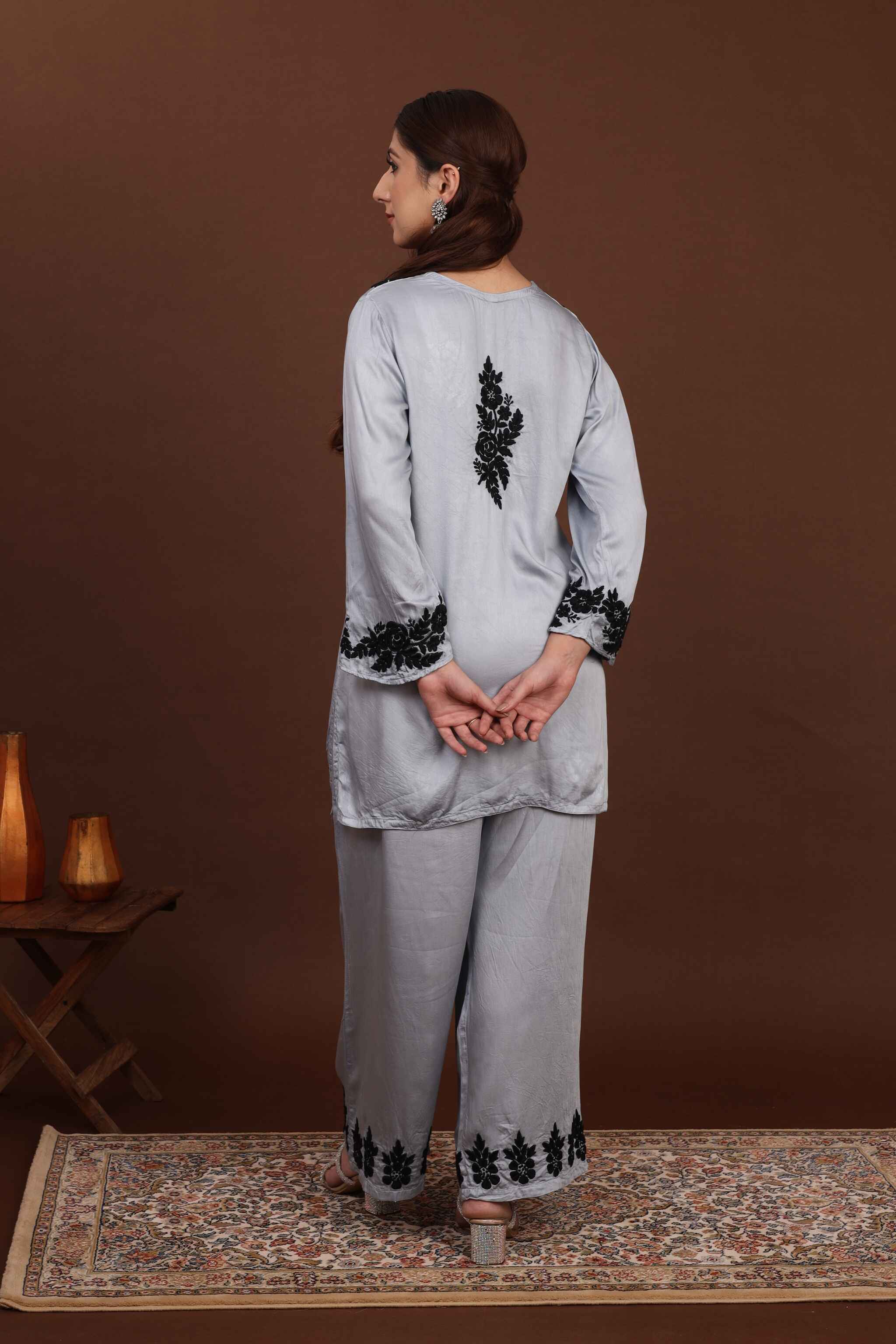 Back view of chikankari co ord set of Woman wearing a light gray traditional outfit with black embroidery on a brown background