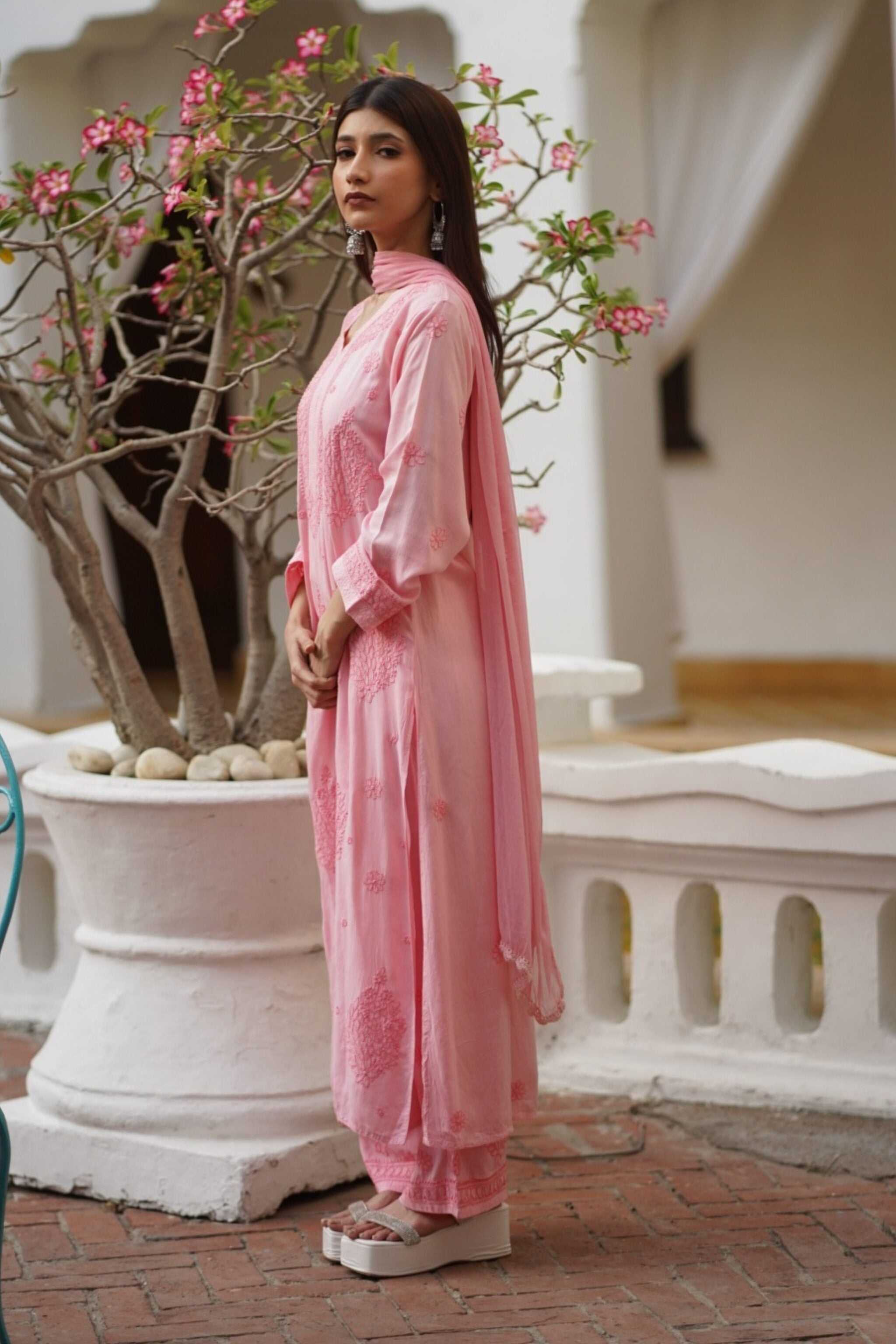 Woman in a pink traditional chikankari kurta set standing next to a potted plant with pink flowers.