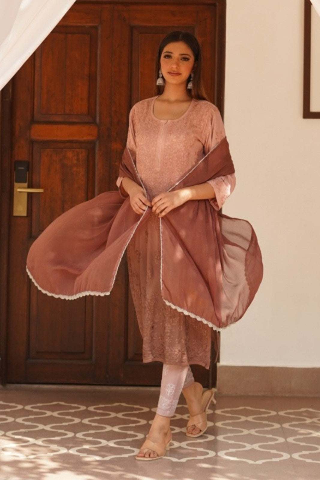 Woman in a pink traditional chikankari kurta set with a brown dupatta standing in a room with a wooden door.