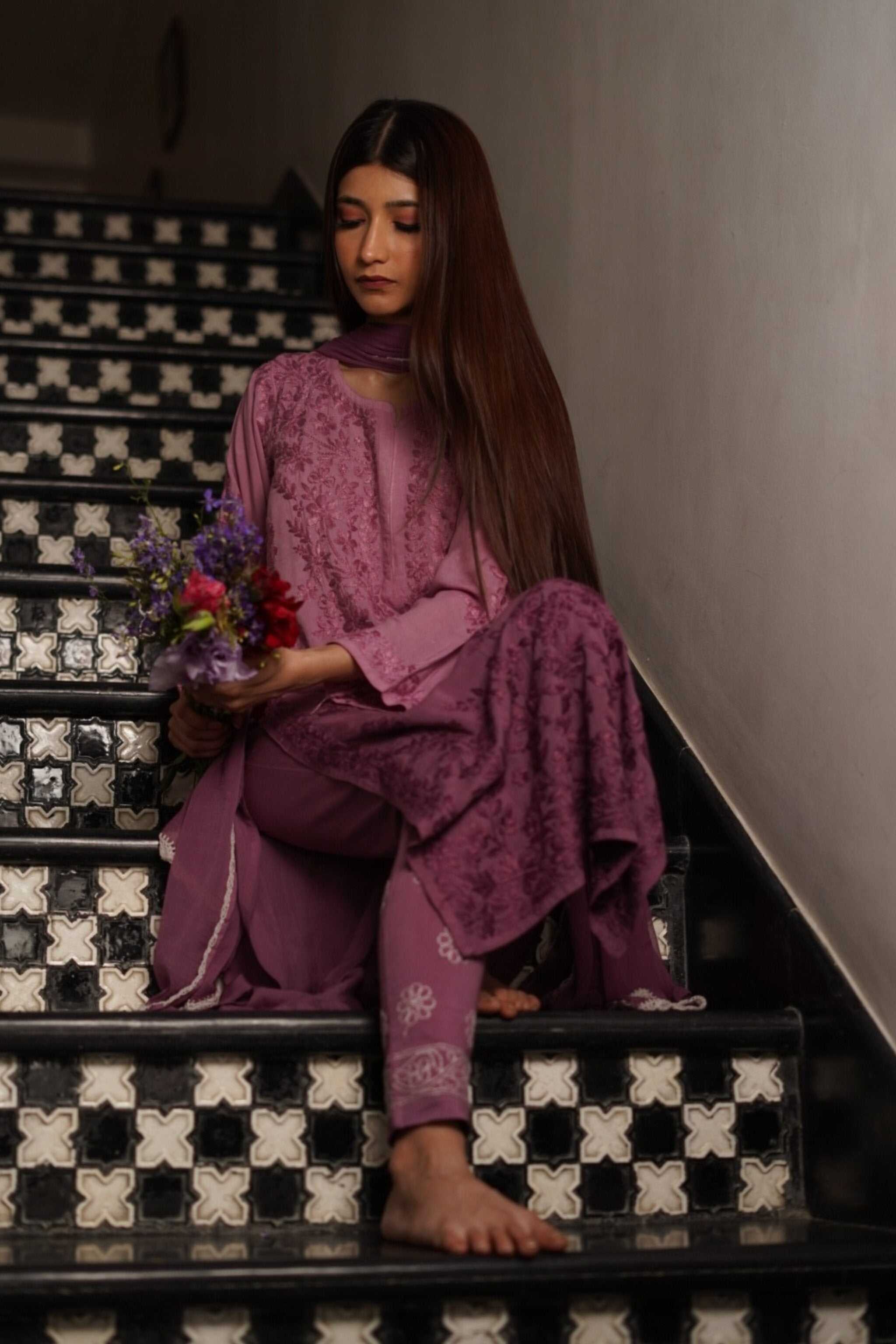 Woman in a purple chikankari suit sitting on a black and white patterned staircase holding flowers.