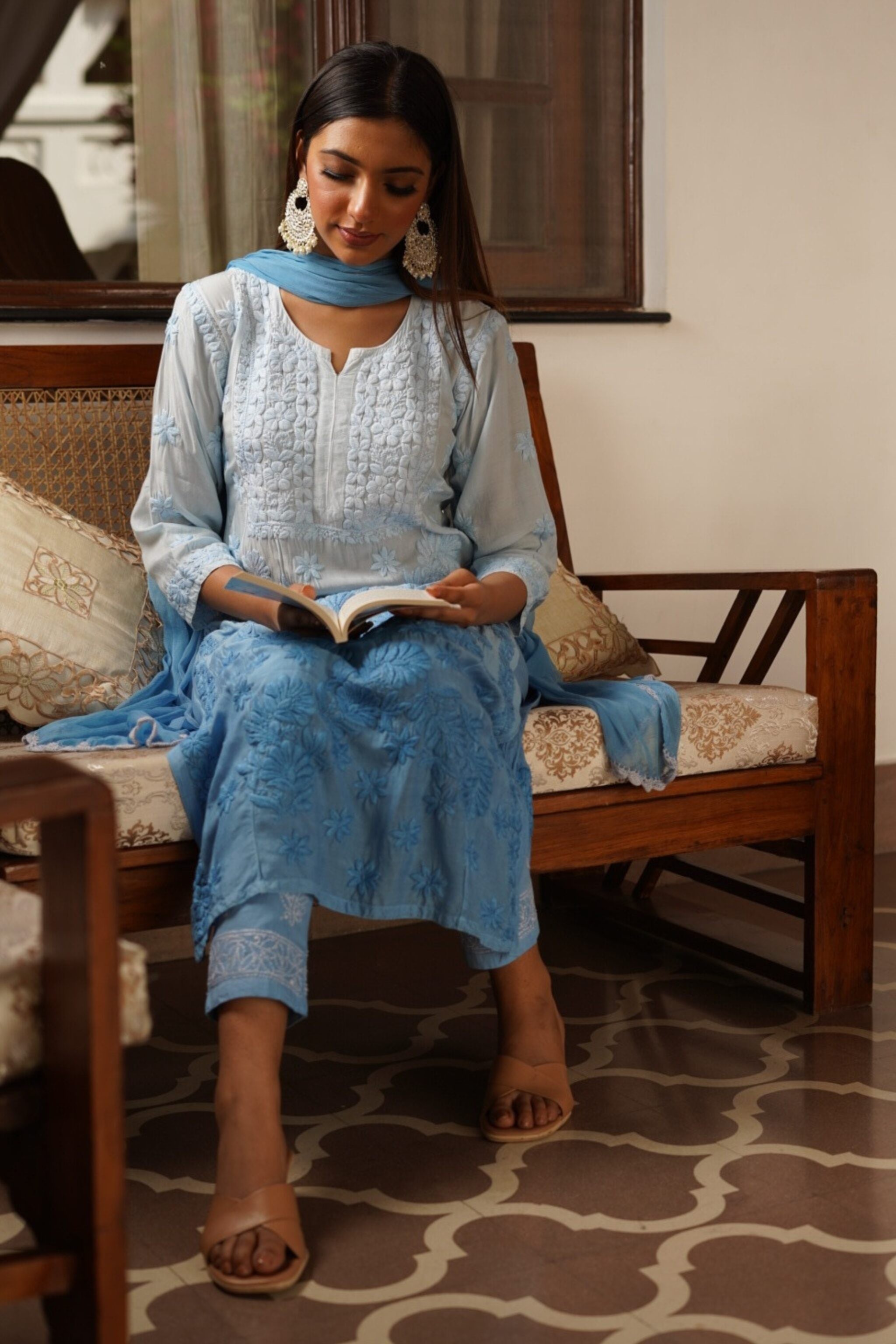 Woman in a blue traditional chikan kurta set reading a book on a wooden bench.
