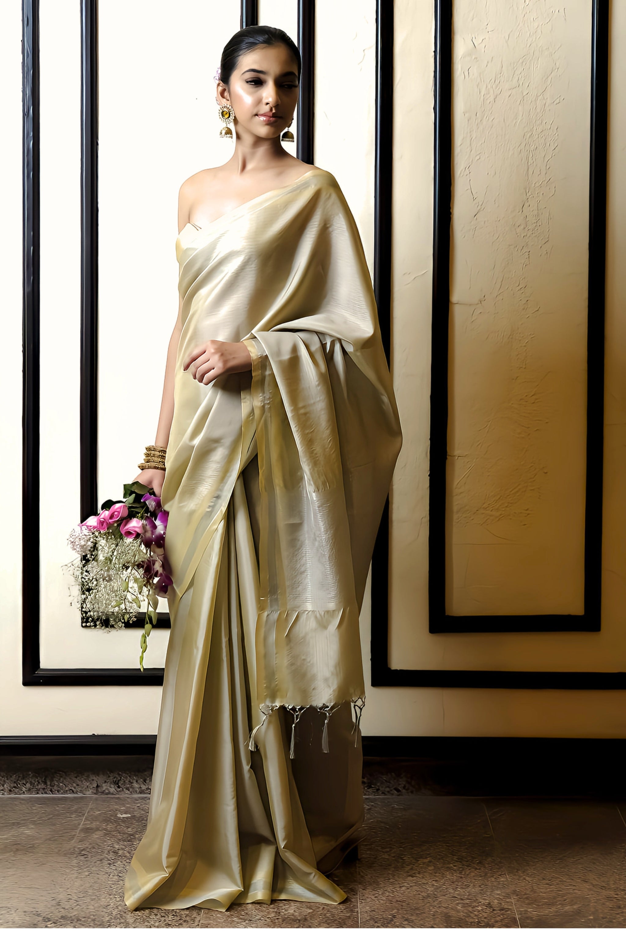 A woman drape the Riwaaz Sheen Gold Tissue Banarasi silk saree and standing next to the wall