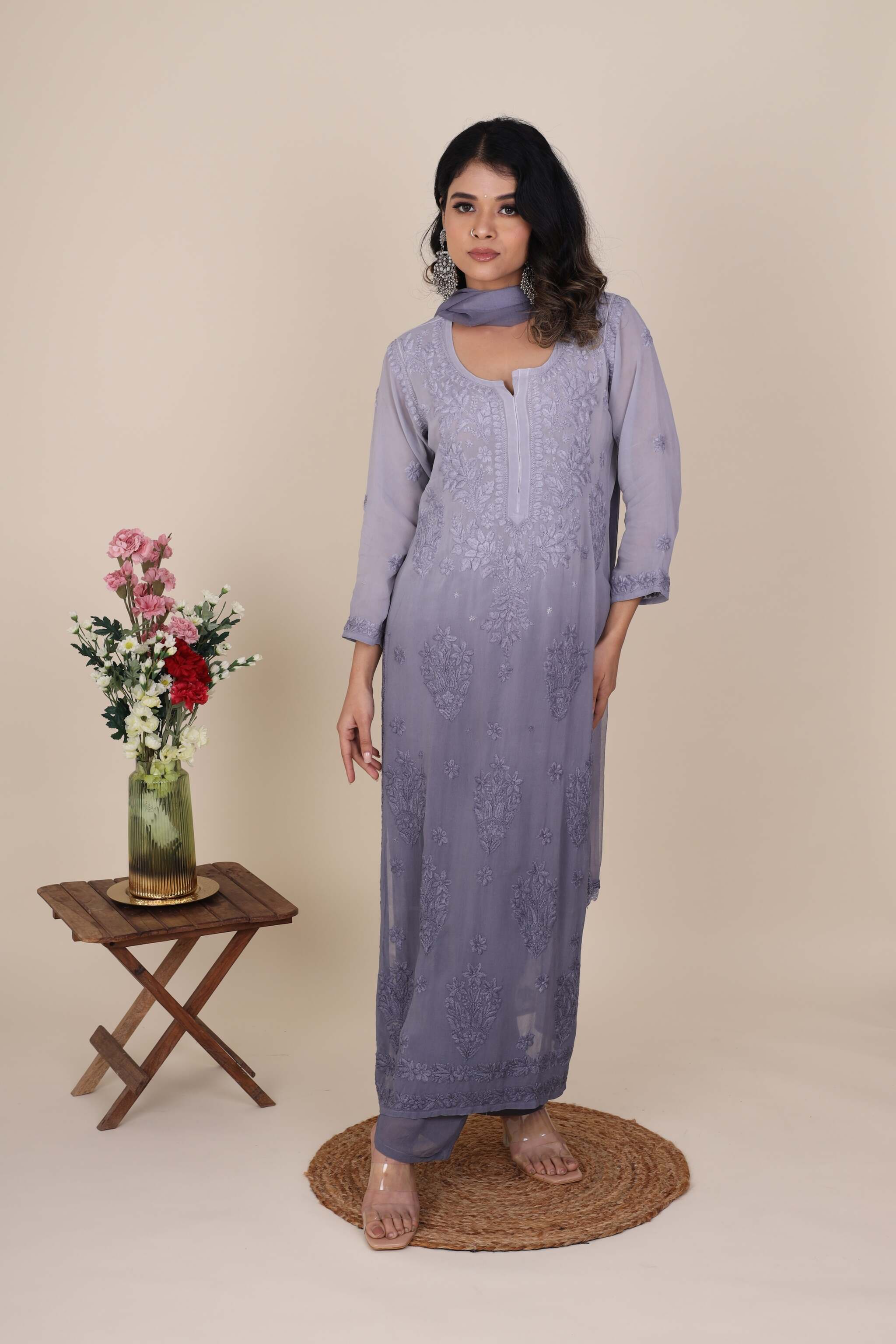 Woman wearing a long grey ombre Heer Agate Chikankari Kurta Set standing next to a small wooden table with flowers on a beige background