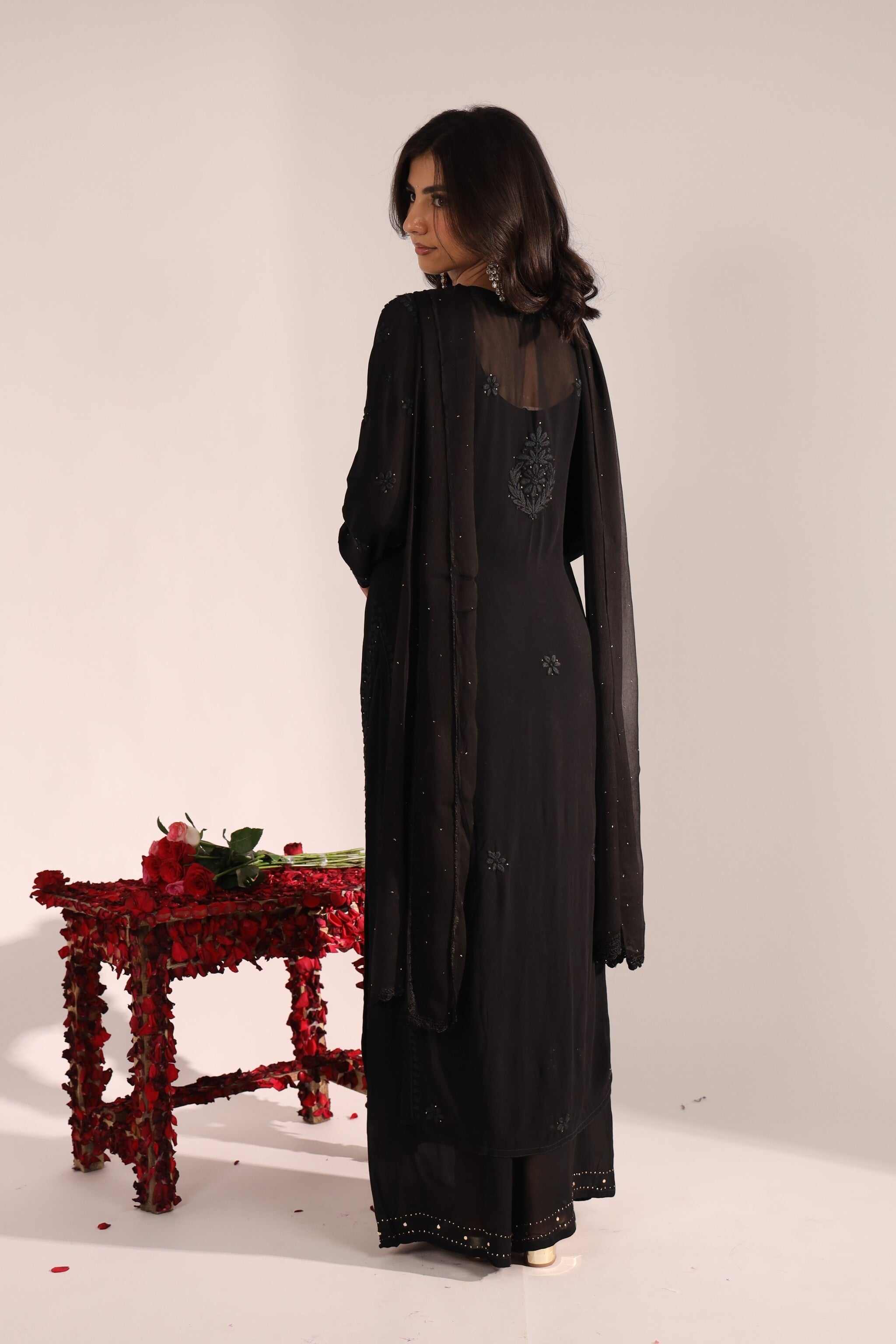 Woman wearing a long black suit with floral embellishments in a minimalistic setting.