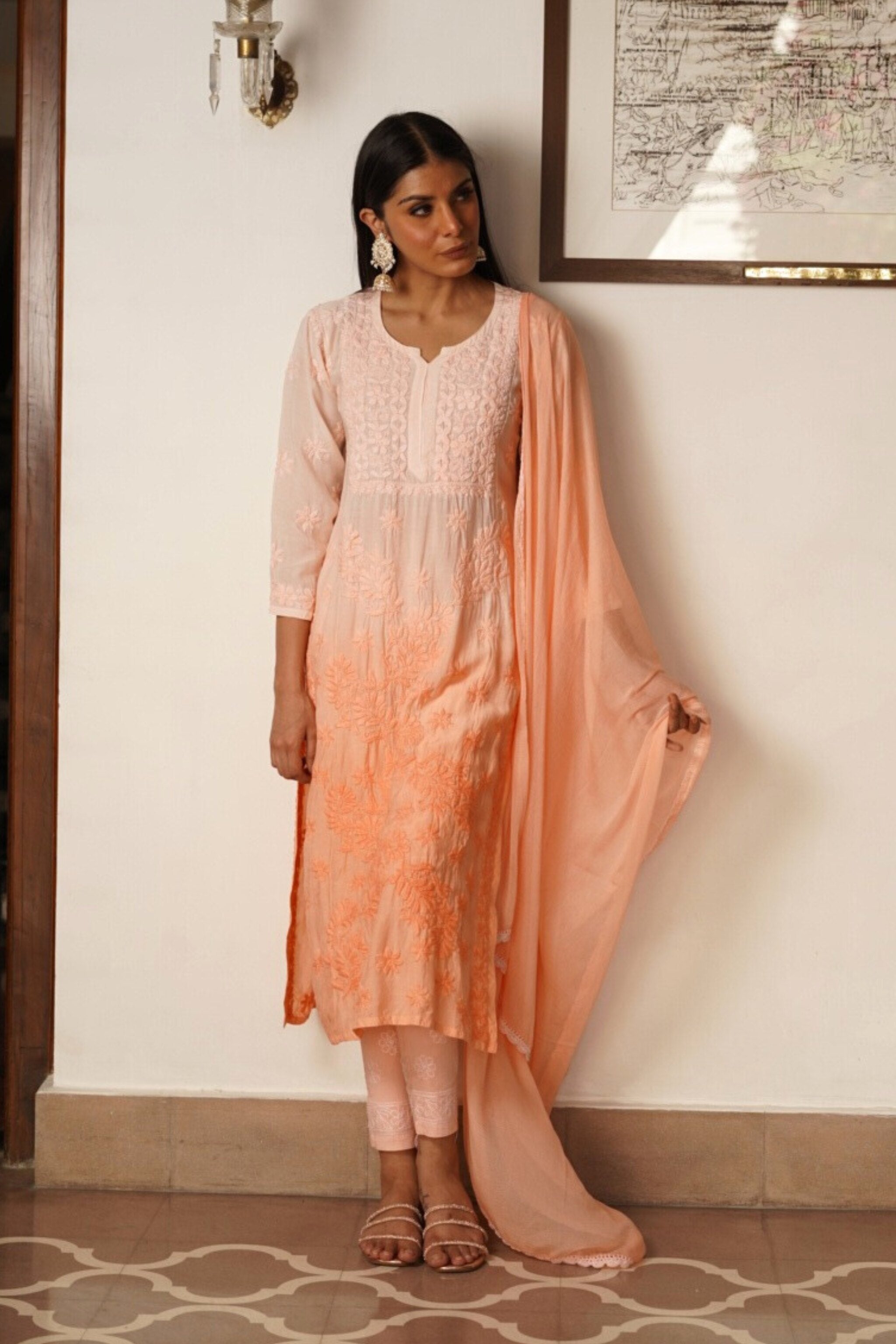 Woman in peach traditional chikankari kurta set standing against a white wall.