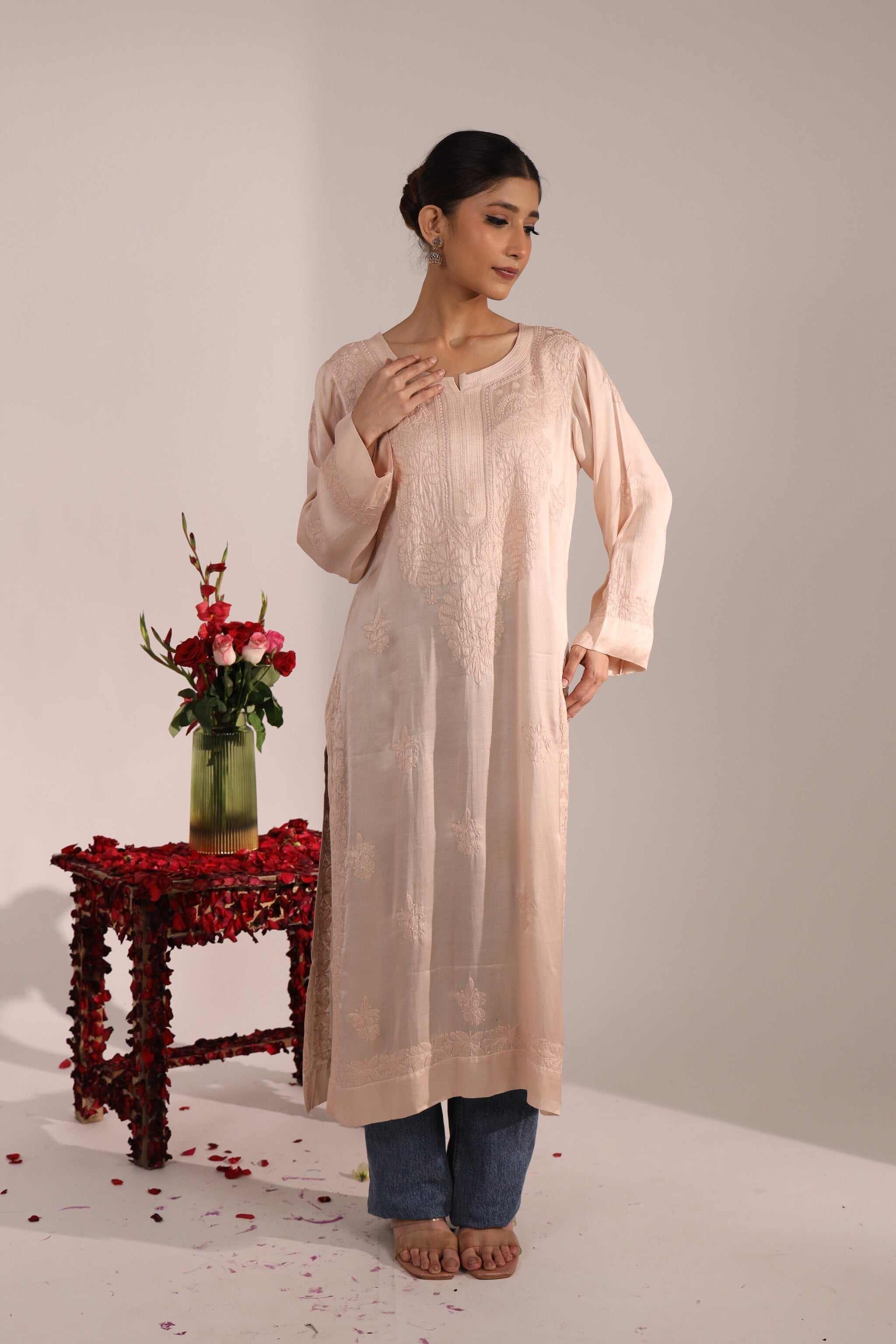 Woman wearing pastel pink Chikan Kurti paired with jeans, standing near a wooden table with flowers – elegant Indo-western look.
