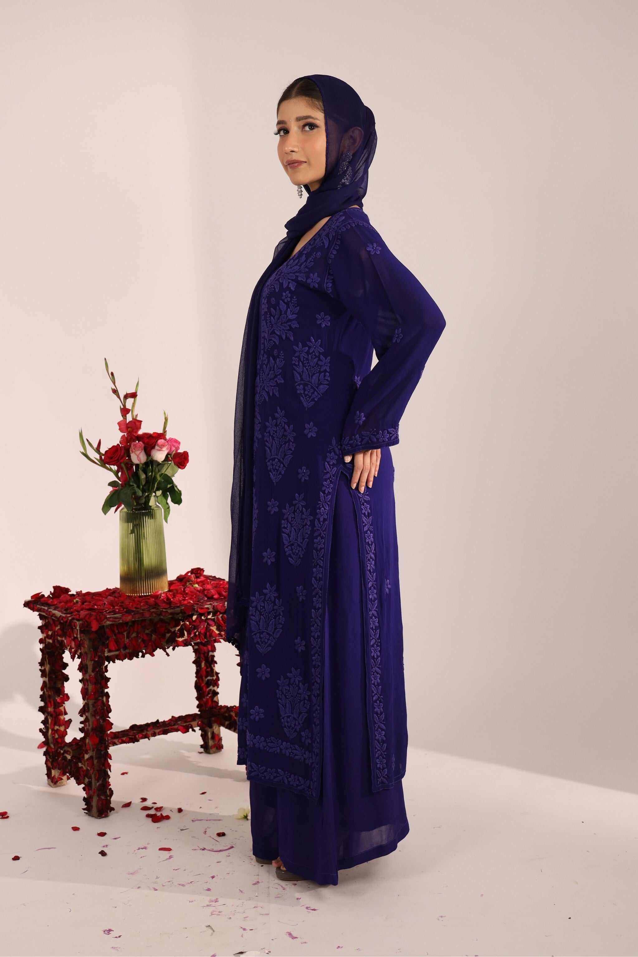 Woman in a blue embroidered Nayantara chikan suit standing next to a table with flowers on a white background