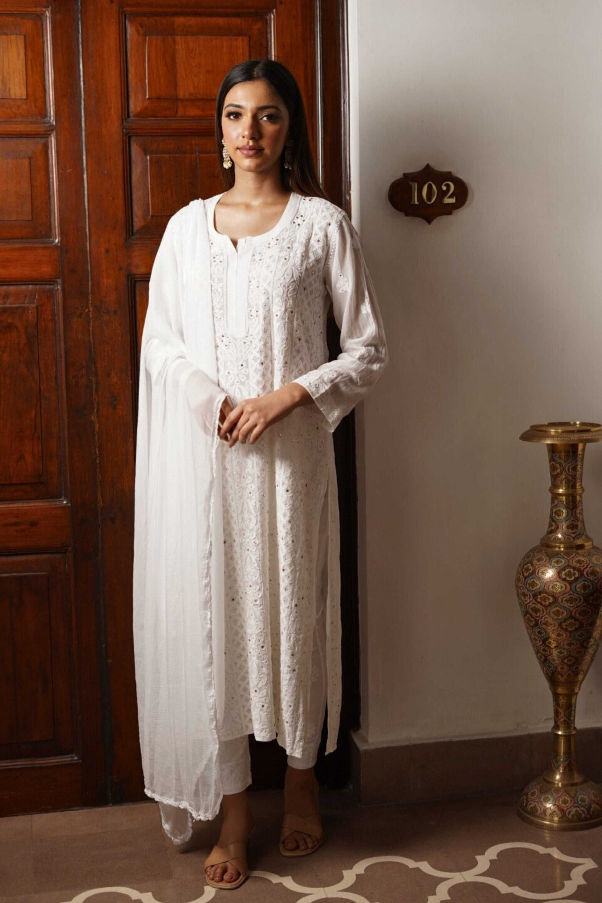 Woman in a white chikankari suit standing in a room with wooden paneling and a decorative vase.